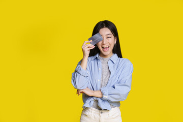 Excited asian girl smiling cover her with bank card right eye ready to spend money. Asian girl in blue shirt with mockup bank card isolated on yellow background. Shopping concept