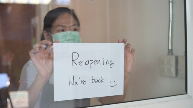 Young Asian business owner coffee shop manager wearing face surgical mask attaching reopen sign for announcing shop reopening after be closed due to business crisis from coronavirus outbreak problem.