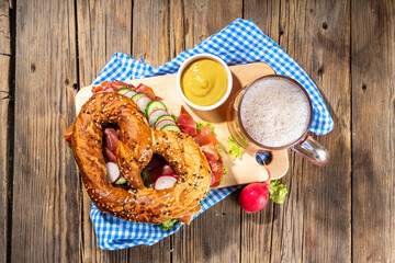 Oktoberfest pretzel bagel sandwich. Oktoberfest day breakfast, burger recipe for holiday beer party. Big pretzel with ham, cheese, fresh vegetables, with radish, beer, checkered tablecloth copy space