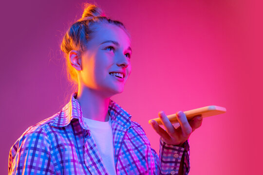 Portrait of adorable young girl, student wearing plaid shirt using cellphone isolated on magenta color background in neon light. Concept of beauty, art, fashion, emotions