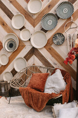 Fototapeta na wymiar A wicker eco chair with an orange plaid, and an orange and white pillow stand on a white carpet. In the background is a wooden wall decorated with white and black plates.