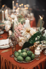 Wedding banquet table decor with orange tablecloth. In the foreground are lime fruits on a tray, white candles in candlesticks, black glasses. In the background are white flowers, candles, green leave