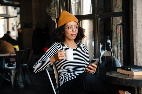 Young African American Woman Drinking Tea And Using Cellphone In Cafe