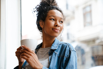 Black young woman smiling and drinking coffee by window in cafe