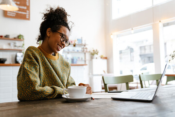 Black young woman working with laptop while sitting in cafe