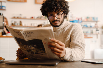 Indian young curly man wearing eyeglasses reading newspaper in cafe
