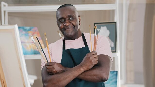 Portrait African male artist holding brush for drawing happy old 50s painter man senior middle-aged talented designer crossing hands looking at camera in art studio posing enjoying painting pictures