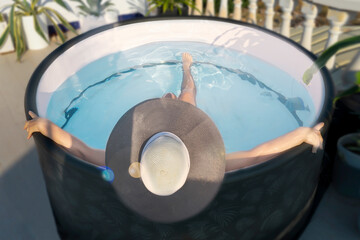 woman in a hat bathes in a small mini-pool with a jacuzzi with blue water on the roof of the house in the sun against the backdrop of tropical plants, top view - obrazy, fototapety, plakaty