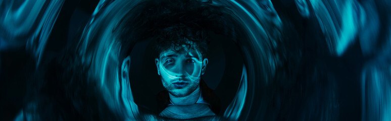 curly man looking at camera through futuristic neon blue circle on dark background, banner.