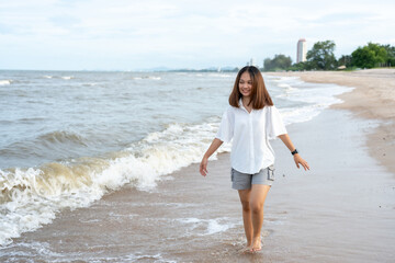 A young tourist, a long-haired Asian woman in a white shirt, is strolling along the beach, where the waves crash with a cheerful expression on her holiday. evening atmosphere sunset