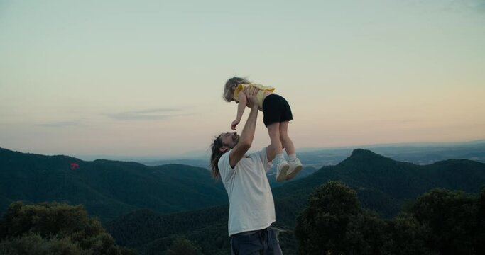 Young adult man playing with toddler girl on epic mountain view. Cheerful family together on summertime trip adventure. Father tosses his daughter over himself at sunset and smile