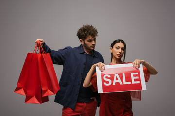 couple in trendy outfits holding shopping bags and paper with sale lettering isolated on grey.