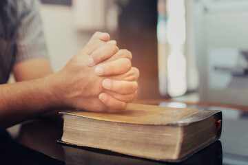 Close up of a man hands praying on the open holy bible on a table indoor with the windows light lay...