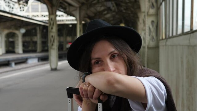 A young tourist woman in a hat with a suitcase at the train station on a summer day. A tourist is resting from a long walk in a new city.