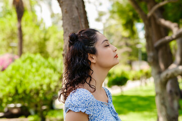 Side view of cute millennial woman wearing a blue floral summer dress standing on city park, outdoors breathing fresh air. Concept of healthy lifestyle.