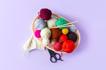Red, yellow, white, purple, brown, green balls of thread in a beige basket with knitting needles made of bamboo and scissors on a lilac background, flat lay