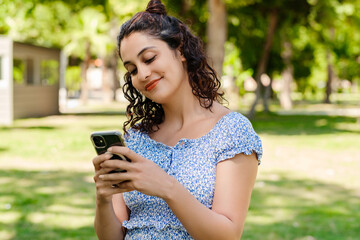 Young woman smiling confident wearing summer dress standing on city park, outdoors looking at the phone screen and using phone. Messaging with friends, watching video or scrolling on social media.