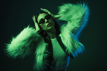 stylish young woman in faux fur jacket and sunglasses posing on blue.