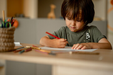 A three-year-old boy with long dark hair sits at home at a table and draws with colored pencils. Child and creativity