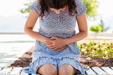 Young beautiful woman wearing summer dress on city park, outdoors with hand on stomach because indigestion, painful illness feeling unwell. Ache or pregnancy concept.