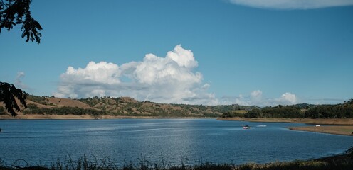 Scenic view of a blue river in a rural area surrounded by small hills in cloudy sky background