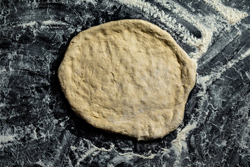 Pizza base from dough on a dark table top view.