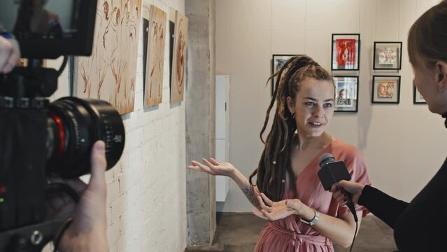 Over-shoulder of young Caucasian female artist with dreadlocks giving interview to journalist, unrecognizable cameraman filming in gallery on exhibition opening