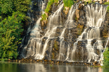 Fototapeta na wymiar Artificial waterfall above river in summer day. Water cascade flowing on stones. Splash stream between the trees. Cold current descends on the shore. City recreation park.