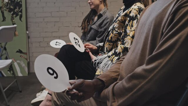 Midsection side-view of cropped buyers sitting on chairs indoors on auction, holding bid cards with numbers