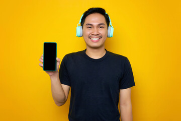 Smiling young Asian man in casual t-shirt with headphones, showing mobile phone with blank screen isolated on yellow background