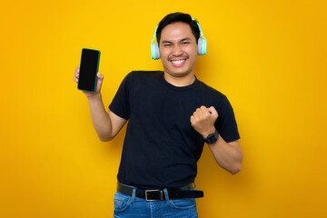 Excited young Asian man in casual t-shirt with headphones, showing mobile phone with blank screen, making winner gesture isolated on yellow background