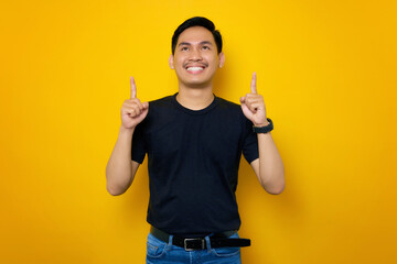 Excited young Asian man in casual t-shirt pointing fingers up at copy space isolated on yellow background. People lifestyle concept