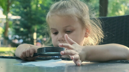 Little girl carefully looks into the lens at the salt. Close-up of blonde girl is studying salt crystals while looking at her through magnifying glass while sitting in street cafe in the park.