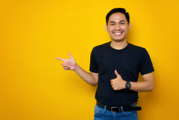 Cheerful young Asian man in casual t-shirt pointing finger aside at copy space, showing thumb up isolated on yellow background. Great offer concept