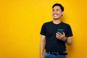 Smiling young Asian man in casual t-shirt holding mobile phone and looking at aside isolated on yellow background. People lifestyle concept