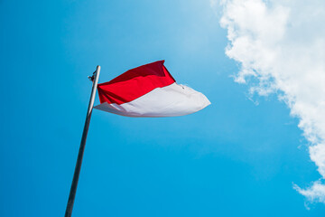 This red and white flag is the national flag of the Indonesian state which is flying in a clear blue sky with clumps of clouds around it