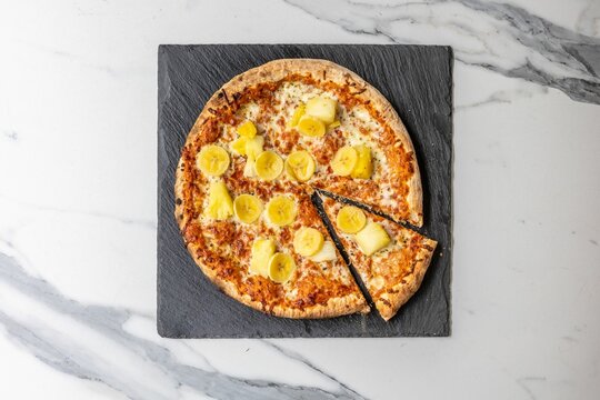 Shot Of A Pizza With A Slice Cut Out Topped With Bananas And Pineapple On A Marble Background