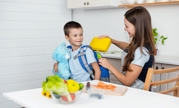 Caring mother puts yellow plastic lunch box to her son in a school backpack. School food or lunch, concept image.