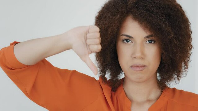 Headshot portrait indoor displeased millennial woman with curly hair looking at camera showing thumbs down rejection finger dislike. Dissatisfied girl disapproval disappointment face reaction bad idea