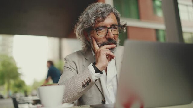 Mature businessman with beard in eyeglasses wearing gray jacket sits on cafe. Middle aged manager successful man user on laptop pc computer sit at cafe outdoors
