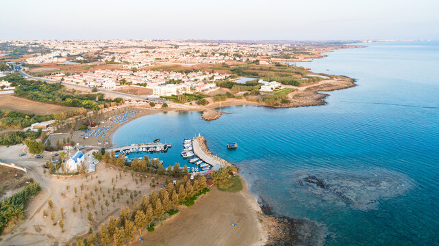 Aerial bird's eye view of coastline sunset, landmark white washed chapel Agia Triada beach, Protaras, Famagusta, Cyprus from above. Tourist attraction Ayia Trias bay church, sand, port at sunrise.