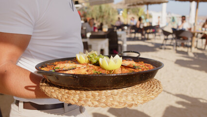 Professional waiter bringing different plates - paella, salad, prawns in tempura - to a family's table in a restaurant on the beach