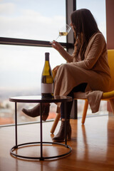 woman sitting on the chair near low table with bottle of wine and drinking white wine and looking...