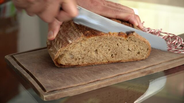 Hand slicing fresh organic artisan bread with knife on wooden board. Healthy food, eat, cook, bakery