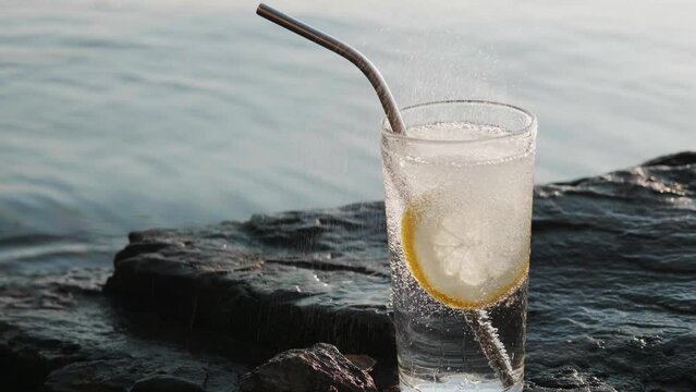 Round slice of lemon falls into glassful of soda water, drink in transparent tall glass stands on stony seashore under the bright sun.