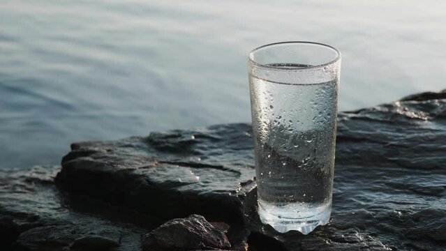 Non-alcoholic drink in transparent tall glass stands on stony seashore under the bright sun.