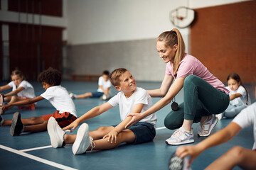 Happy boy stretching on floor with help of PE teacher during exercise class at school gym.