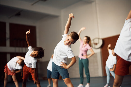 Happy schoolboy doing stretching exercise during physical education class at school gym and looking at camera.