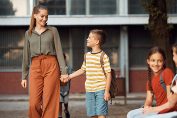 Happy schoolboy and his mother holding hands while walking through schoolyard.