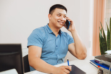 a young Asian businessman is talking on the phone to discuss work in the office. the man is Asian.